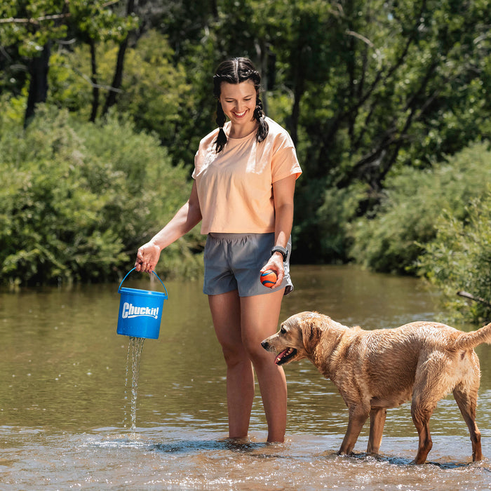 A woman stands in a creek holding the Chuckit! Bucket With Ultra Balls by Chuckit!, smiling at her wet golden retriever. Lush green trees frame their playtime with this favorite fetch toy.
