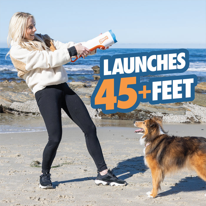 A woman on a beach uses the Chuckit! Cannon & Cannon Max Dog Ball Launcher by Chuckit! to play fetch with her brown and white dog. Bold text beside them reads, Launches 45+ feet. The ocean and rocks are in the background.