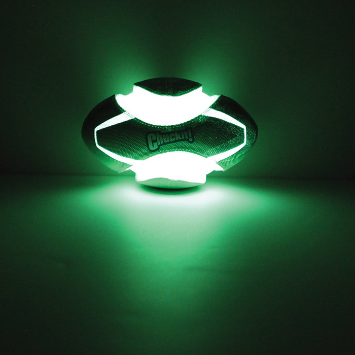 A glowing green Chuckit! Fumble Fetch Max Glow football with illuminated panels rests on a dark surface, casting bright green light. The word Chuckit! is faintly visible on this glow-in-the-dark fetch toy.