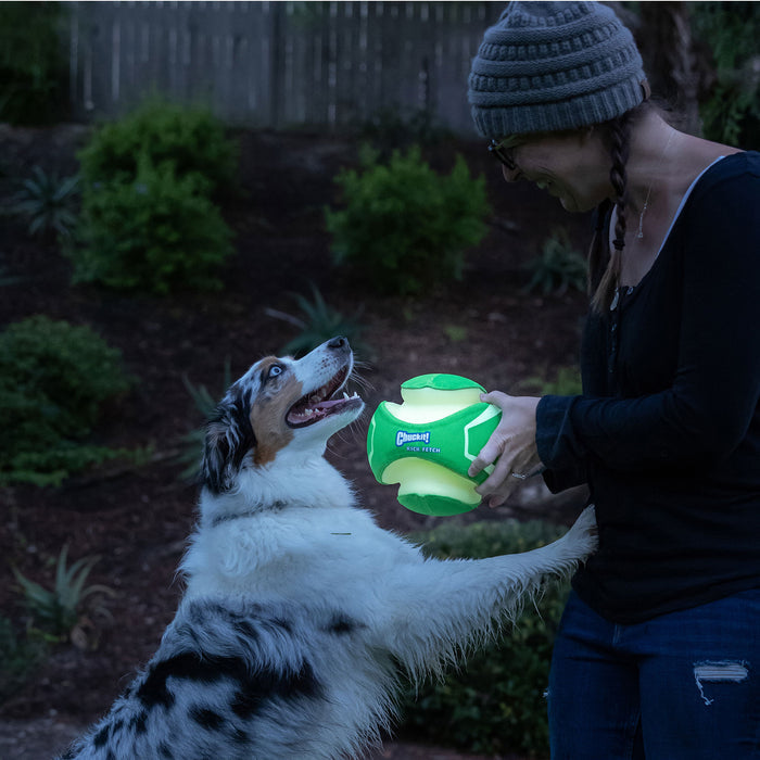 A woman in glasses and a beanie smiles as she holds a Chuckit! Kick Fetch Max Glow toy from Chuckit!, while her happy dog stands on its hind legs, reaching for the glowing ball in a garden.