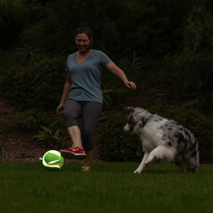 A woman in athletic wear kicks a Chuckit! Kick Fetch Max Glow ball by Chuckit! on the grass, while a black and white dog excitedly chases it outdoors amid lush greenery.
