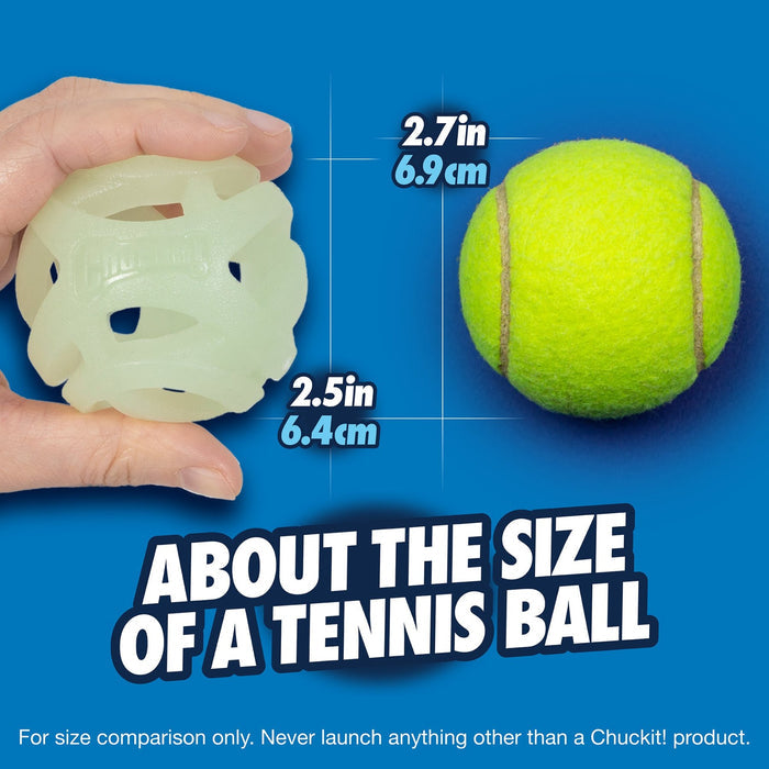 A hand holds a Chuckit! Max Glow Air Fetch Ball by Chuckit! (2.5 in/6.4 cm) next to a yellow tennis ball (2.7 in/6.9 cm). Text: "About the size of a tennis ball." The Max Glow Air Fetch Ball is white and breathable.