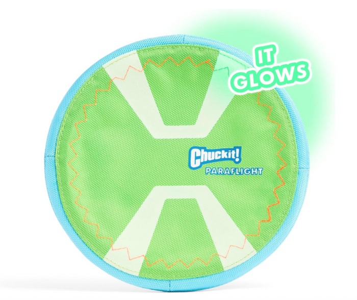 A green and blue Chuckit! Paraflight Max Glow Flyer Dog Toy with stitched edges glows at the corner. Text reads “IT GLOWS.” This Chuckit! fetch toy is perfect for nighttime play with your dog.