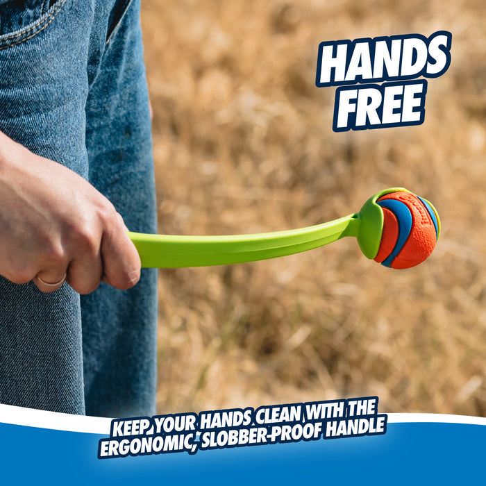 A person holds a green Chuckit! Sport Launcher with an orange and blue tennis ball outdoors on grass. Text: HANDS FREE—Keep your hands clean with the ergonomic Chuckit! handle for easy, hands-free pickup.