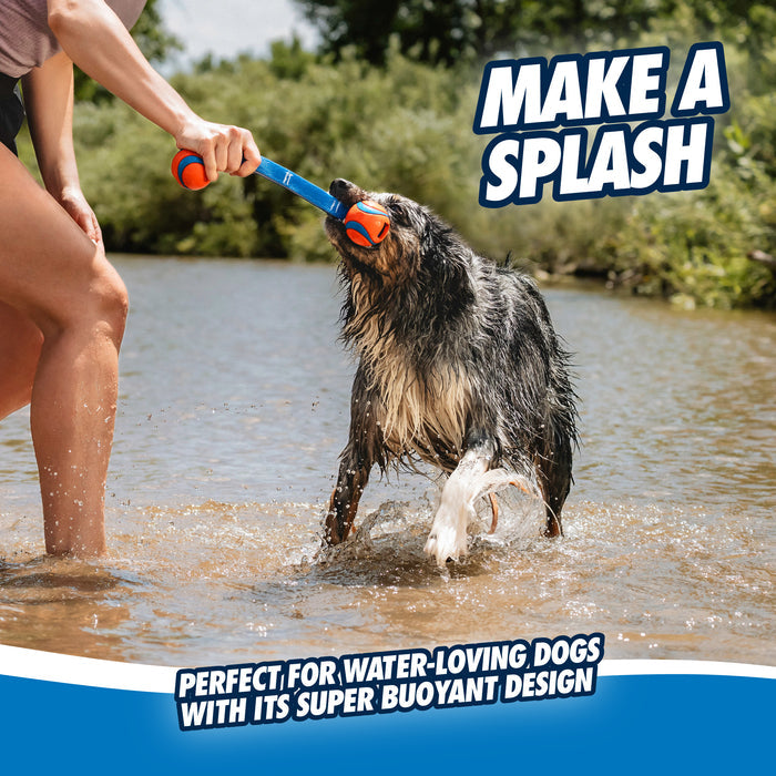 A wet dog stands in shallow water, grabbing the Chuckit! Ultra Duo Tug Dog Toy Med by Chuckit! Text reads: MAKE A SPLASH—perfect for water-loving dogs with its super buoyant design. Trees and sky fill the background.