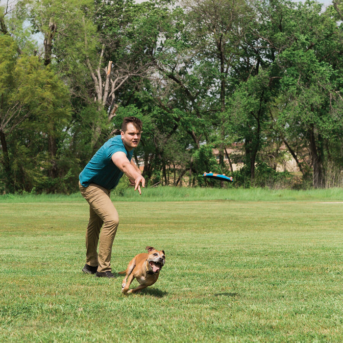 A man in a teal shirt and khaki pants throws a Chuckit! Whistle Flight Dog Fetch Toy by Chuckit! across a grassy field as his brown and white dog eagerly runs ahead to catch it. Trees fill the background.