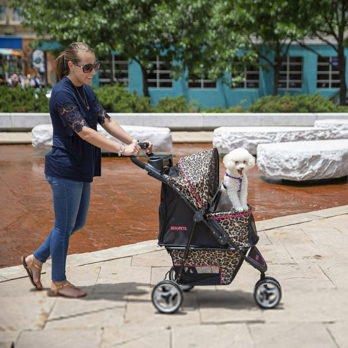 Wearing sunglasses, a navy blouse, and jeans, a woman strolls outdoors with trees and a blue building behind her, pushing a relaxed small white dog in the Gen7Pets Cheetah Promenade Pet Stroller by Gen7, featuring a smart-comfort pad.