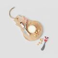 A styled flat-lay of a wooden cheese board with a round cheese wheel, two decorative cheese knives, peanuts, and candy canes on a white marble surface over printed papers.