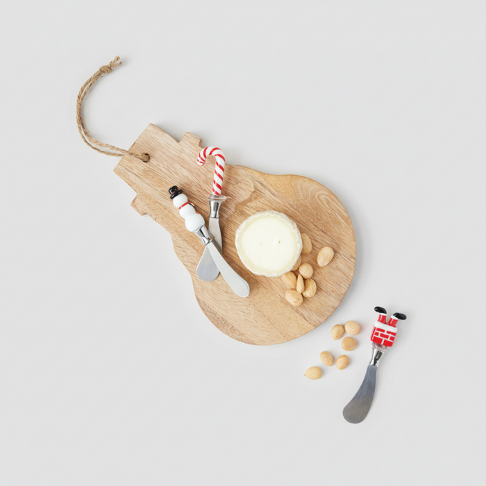 A styled flat-lay of a wooden cheese board with a round cheese wheel, two decorative cheese knives, peanuts, and candy canes on a white marble surface over printed papers.