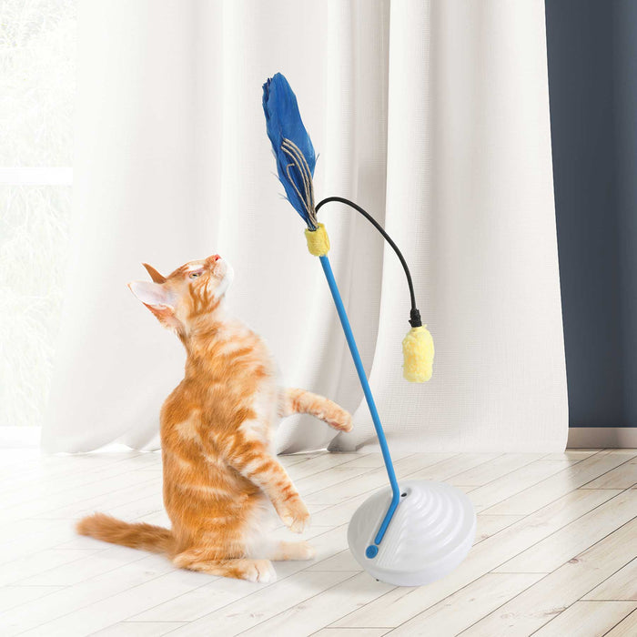 An orange and white cat stands on its hind legs, reaching for the JW Catnip Mister by JW Pet, a blue interactive cat toy attached to a flexible rod on a white base, with white curtains and a light wood floor in the background.
