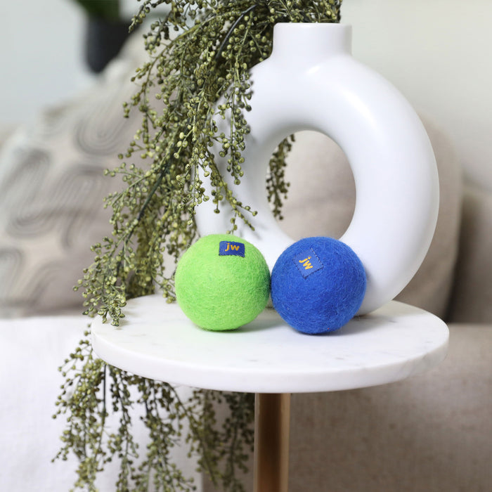 A green and a blue JW Wool-ee Ball dog toy by JW Pet sit on a round white marble table by a modern white vase with greenery in a cozy living room.