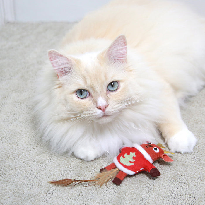 A fluffy cream-colored cat with blue eyes lounges on a carpet, playfully batting at a Mad Cat Ugly Sweater Stuffer Reindeer toy, complete with antlers and a feathered tail.