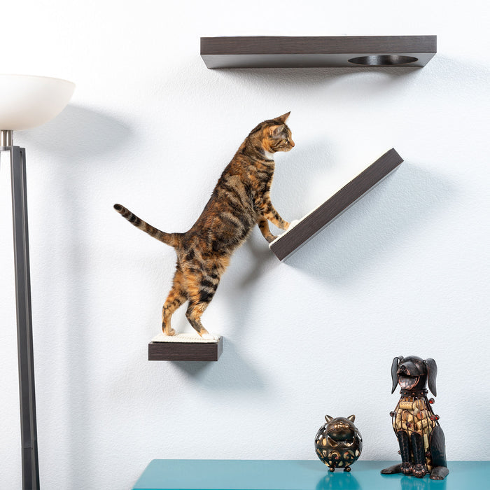 A cat adeptly climbs the PetFusion Wall Activity Shelves for Cats by Pet Fusion, designed like stairs. Below, a decorative cat and dog figurine sits on a teal table, with a floor lamp to the left against a white wall.