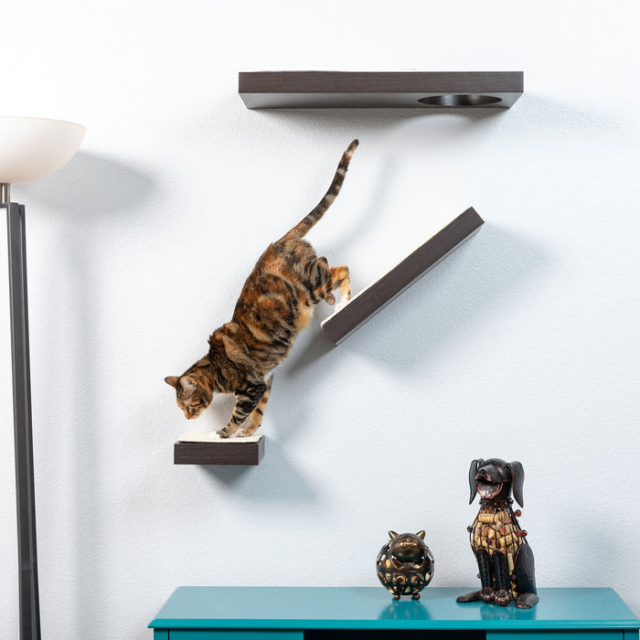 A cat gracefully descends the PetFusion Wall Activity Shelves For Cats by Pet Fusion on a white wall. Below, a teal cabinet displays two decorative figurines—a cat and a dog—while a tall floor lamp stands on the left, completing this brightly lit indoor scene.