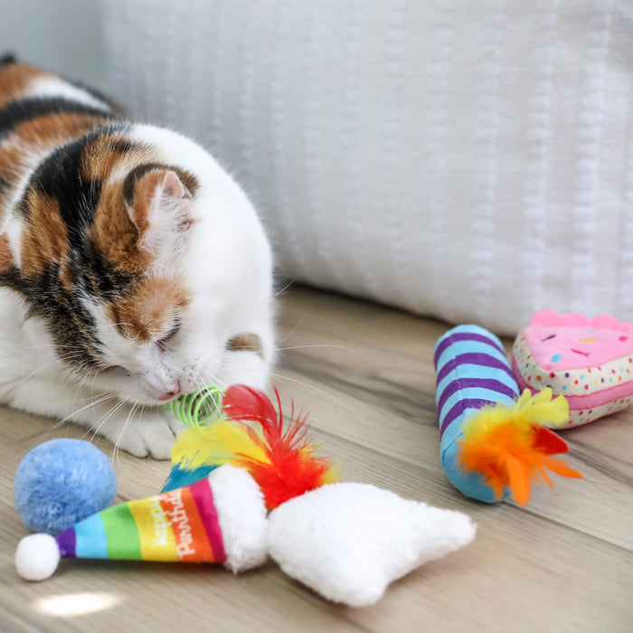 A calico cat plays on a wooden floor with toys from the Quirky Kitty Birthday Box 8pk by Quirky Kitty, including a rainbow party hat, blue pom-pom, and striped feather toy. A white textured cushion appears in the background.
