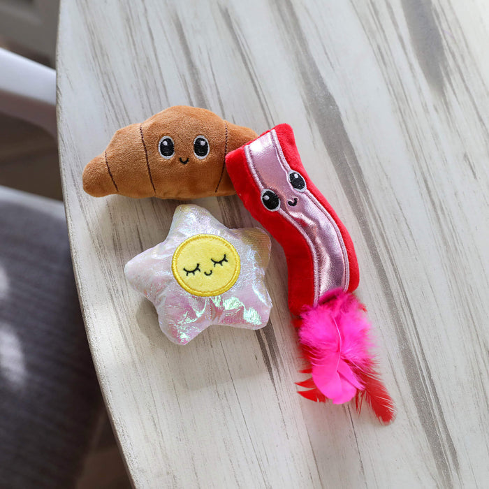 The Quirky Kitty Brunch Bunch 3-Pack Cat Toy Set with Catnip by Quirky Kitty includes a croissant, a bacon strip with a pink feather, and a smiling star—adorable interactive plush cat toys perfect for your feline friend.