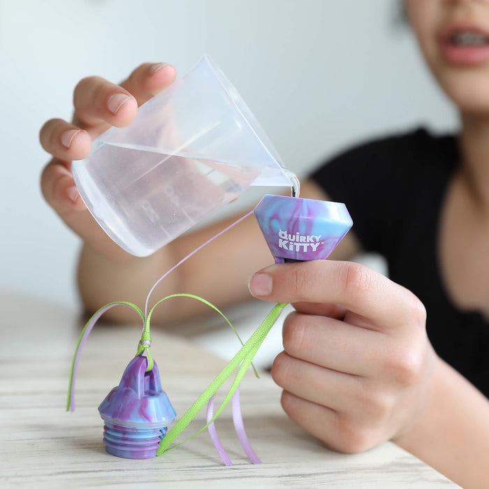 Someone pours water from a clear cup into a colorful Quirky Kitty Chilly Chasers 2pk – Freeze & Play Cat Toy Set container, decorated with ribbons—a playful cat toy from Quirky Kitty—on a light wooden surface.