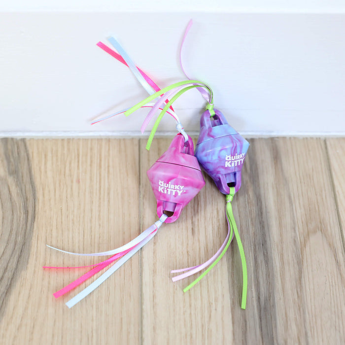 Two Quirky Kitty Chilly Chasers cat toys—a pink and a purple, each with plastic bodies, colorful ribbon tails, and quirky KITTY labels—rest on a wooden floor by a baseboard. Cool freeze & play toys from Quirky Kitty for playful cats.