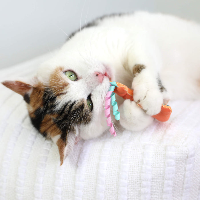 A calico cat with green eyes lies on a white textured blanket, playfully holding and biting the Quirky Kitty Color Changing Shark 2pk – Ocean-Themed Catnip Cat Toys by Quirky Kitty.