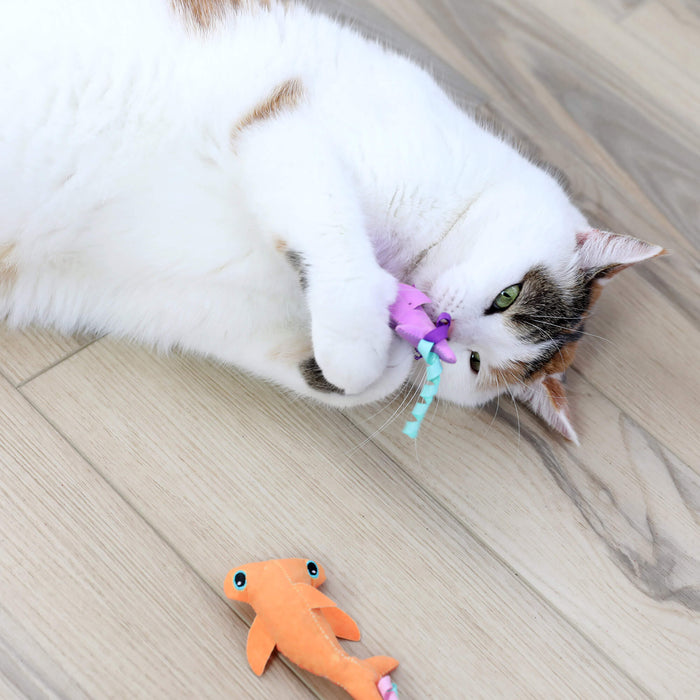 A white cat with brown and black patches lies on a light wooden floor, playing with Quirky Kitty Color Changing Shark catnip toys by Quirky Kitty, while an orange plush fish toy rests nearby.