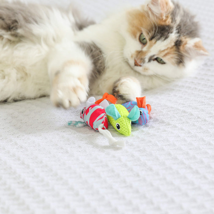 A fluffy cat lounges on a white surface, reaching for the Quirky Kitty Cool Mice 3 Pack—ocean-themed cooling cat toys with catnip—scattered playfully in front of it.