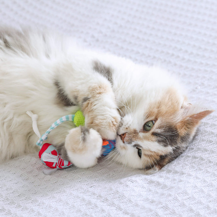 A fluffy white and gray cat lies on a textured white blanket, playfully holding and biting a Quirky Kitty Cool Mice Ocean-Themed Cooling Cat Toy with Catnip.