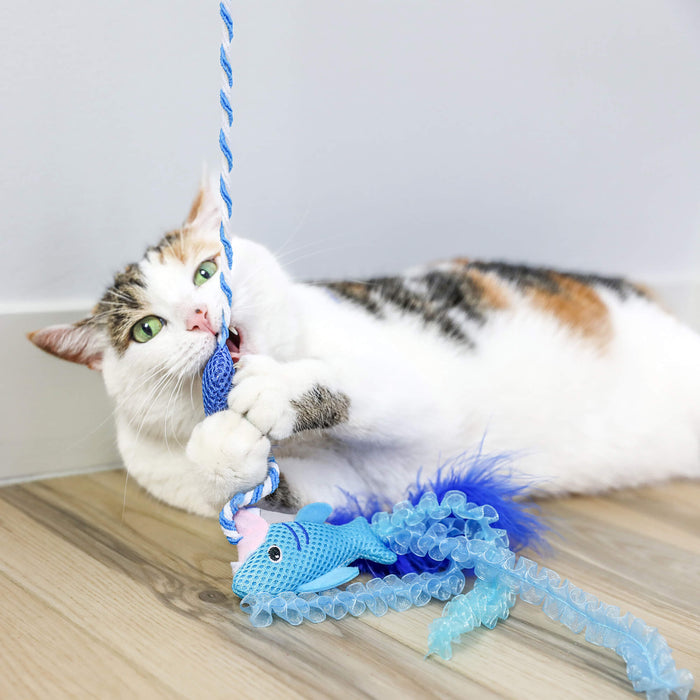 A white and brown cat lies on a wooden floor, playfully biting and holding the Quirky Kitty Cool Shark Wand—an interactive cooling cat toy with catnip from Quirky Kitty—as it looks up with bright green eyes.