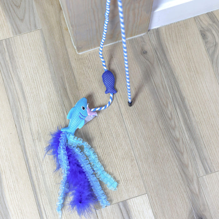 The Quirky Kitty Cool Shark Wand – an interactive cooling cat toy with catnip by Quirky Kitty – features a blue shark on a blue-and-white striped string, lying on a light wood floor next to a wall.