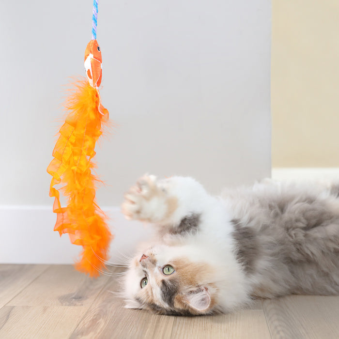 A fluffy cat on a wooden floor playfully reaches for the Quirky Kitty Cute Koi Wand, featuring a lively orange feather and blue rope. The curious felines gaze is set against a pale backdrop with hints of a white wall behind.