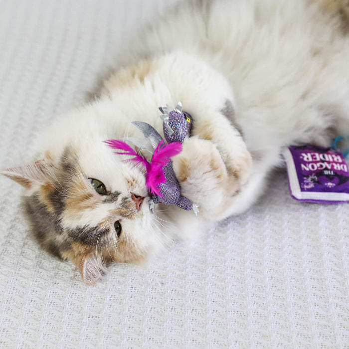 A fluffy calico cat plays on a white surface with the Quirky Kitty Fairy Tail Romance 2pk Cat Toy Set with Catnip by Quirky Kitty. A small purple “Cat Crack Catnip” packet sits beside the feathered toys.