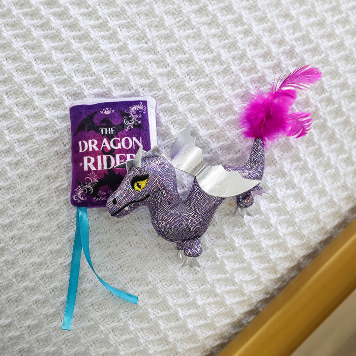 The Quirky Kitty Fairy Tail Romance 2pk Cat Toy Set with Catnip features a plush dragon toy with silver wings and a purple feathered tail next to a purple fabric book, perfect for fairy tale-loving cats.