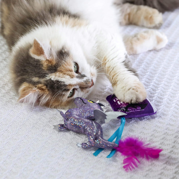 A fluffy, multicolored cat lounges on a white blanket, playing with a Quirky Kitty Fairy Tail Romance Cat Toy by Quirky Kitty—a dragon-shaped toy featuring blue ribbons, a pink feather, and infused with catnip.