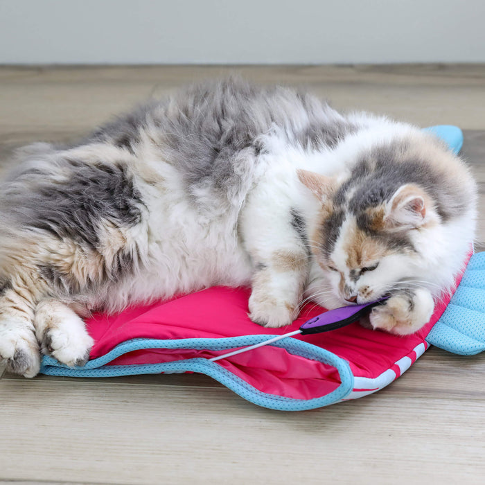 A fluffy gray and white cat relaxes on a wooden floor, lying on its side with its paw and head resting on the Quirky Kitty Fish Cooling Mat – an ocean-themed cat play mat by Quirky Kitty.