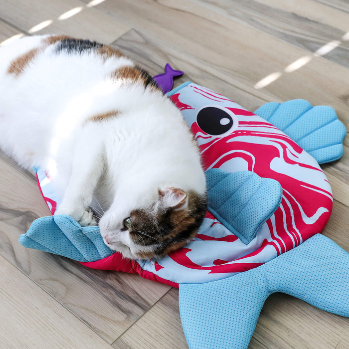 A white and brown cat lies on its side, cuddling with the Quirky Kitty Fish Cooling Mat – an ocean-themed play mat by Quirky Kitty, on a light wooden floor with sunlight casting soft patterns.