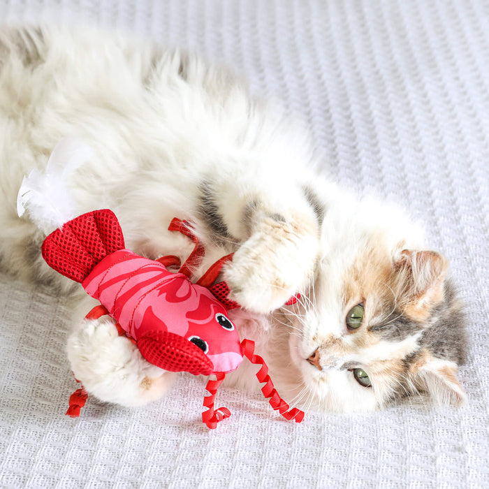 A fluffy calico cat lies on a white textured blanket, playfully batting at the Quirky Kitty Fresh Lobster Kicker—a catnip-filled, ocean-themed kicker toy by Quirky Kitty designed with cooling materials.