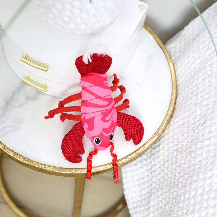 The Quirky Kitty Fresh Lobster Kicker—a catnip-filled, ocean-themed toy—rests on a marble-topped table with gold trim, beside a white textured blanket and stacked white boxes.