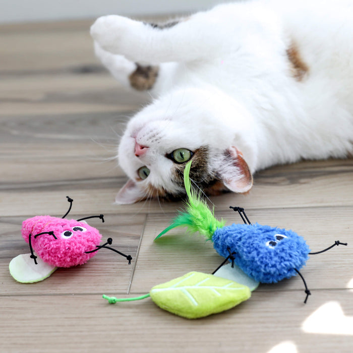 A white and brown cat lies on a wooden floor, looking up at three Quirky Kitty Glow Bugs catnip & silvervine toys—pink bug, blue bug, and green leaf—that glow in the dark.