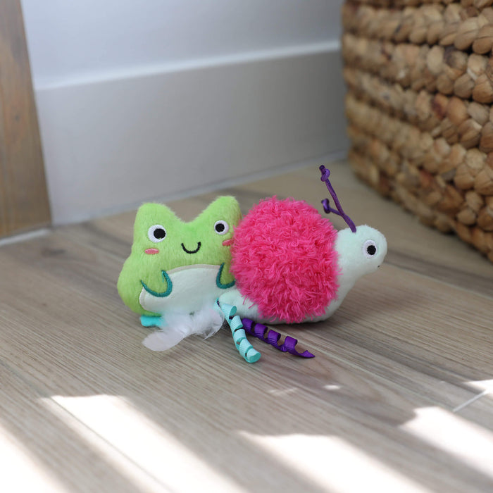 Two plush Quirky Kitty Glow Critters— a green frog and a pink snail—sit on a wooden floor by a basket. Filled with catnip and silvervine, these glow-in-the-dark toys are perfect for playful cats. Available in a convenient 2-pack.