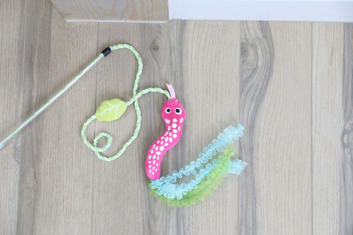 The Quirky Kitty Glow Snake Wand – Glowing Forest Cat Toy with Catnip & Silvervine by Quirky Kitty, featuring googly eyes, ruffled ribbons, and a green leaf, lies on a light wood floor.