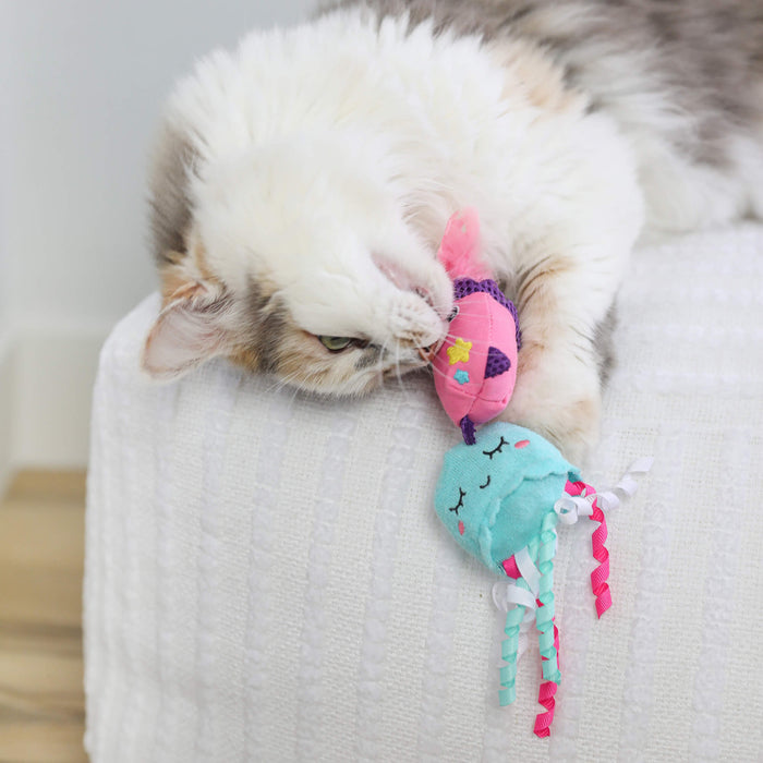 A fluffy white and gray cat plays on a white textured surface, biting a colorful fish toy and the Quirky Kitty Jellyfish cat toy with pink and white ribbon tentacles from the Quirky Kitty Jellyfish & Chill 2-Pack Cat Toys with Catnip.