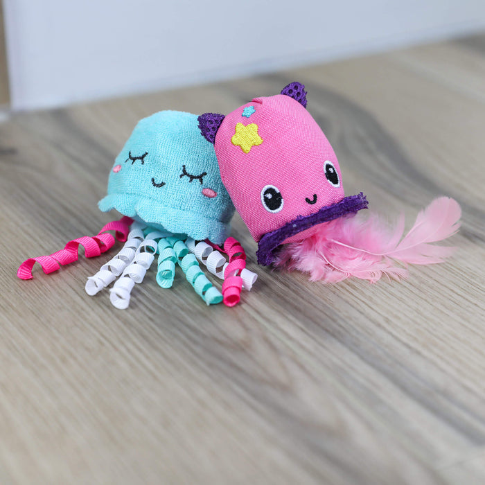 Two Quirky Kitty Jellyfish & Chill 2-Pack Cat Toys with Catnip are on a wooden floor: one is a blue jellyfish with curled ribbon tentacles, and the other is pink with purple trim, cat ears, face details, and pink feathers.