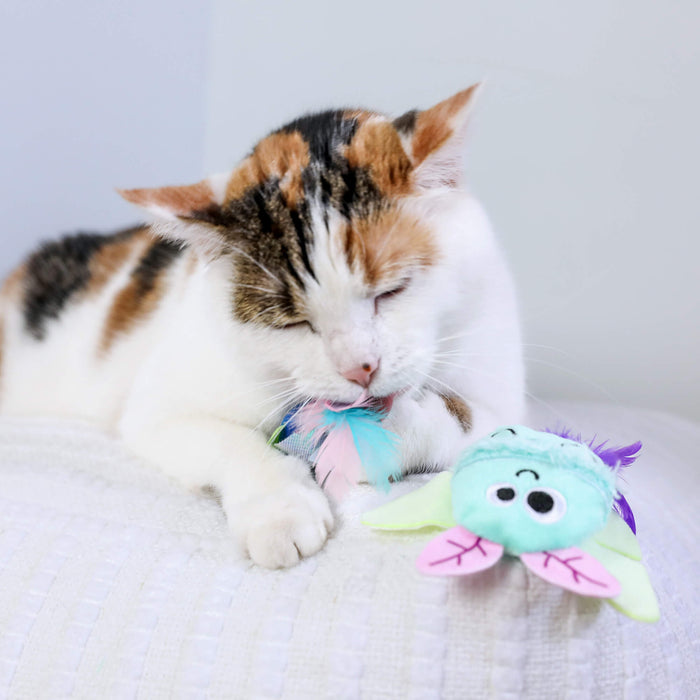 A calico cat relaxes on a white textured surface, playfully biting a Quirky Kitty Luna Bugs Glow-in-the-Dark Cat Toy with feathers, catnip, and silvervine. The cute toy, sold in a 2-pack, features a charming forest-inspired design.