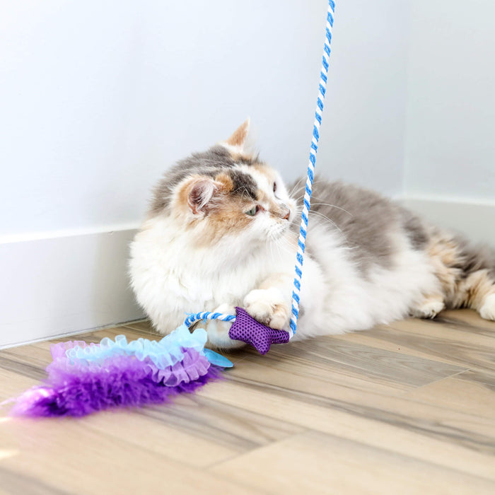 A fluffy calico cat lies on a wooden floor, holding the blue and white rope from the Quirky Kitty Manta Ray Wand interactive toy with catnip, featuring a purple and blue feather, looking away to the left.