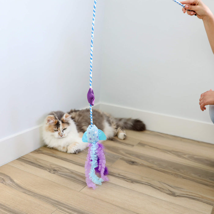 A fluffy white and gray cat lies on a wooden floor near a wall, watching the Quirky Kitty Manta Ray Wand – an interactive cat wand toy with catnip by Quirky Kitty – being held by someone out of frame.
