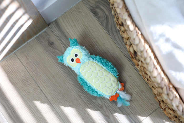 The Quirky Kitty Night Owl Kicker – a turquoise and yellow glow-in-the-dark catnip & silvervine toy shaped like an owl – rests on a light wooden floor near a woven basket and bed, with sunlight casting striped shadows.