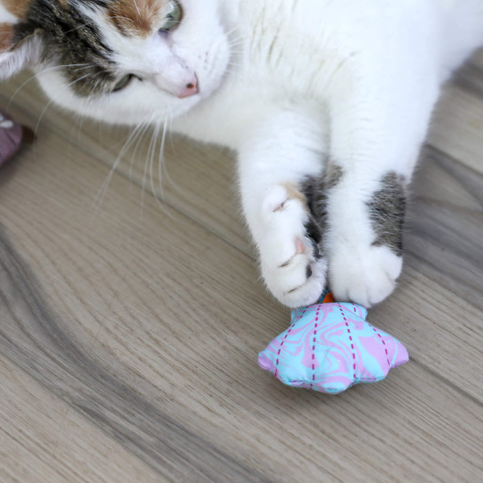A white cat with brown and black markings lies on a wooden floor, playfully holding a Quirky Kitty Otterly Cool ocean-themed cooling cat toy with catnip between its paws.