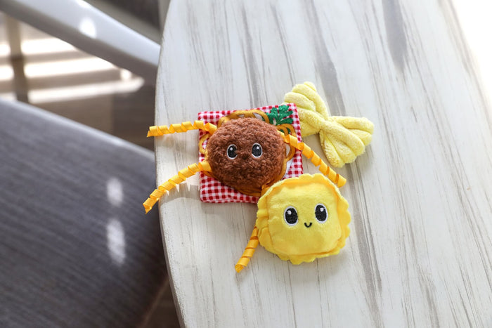 The Quirky Kitty Pasta Pals 3-Pack Cat Toy Set with Catnip features adorable plush cat toys in fun designs, shown here on a wooden table beside a red checkered pillow and yellow plush. A playful set from Quirky Kitty for your home.