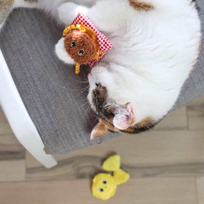 A white and brown cat lounges on a gray chair, playing with a brown plush toy from the Quirky Kitty Pasta Pals 3-Pack Cat Toy Set with Catnip, while the yellow toy from the set lies on the wooden floor below.