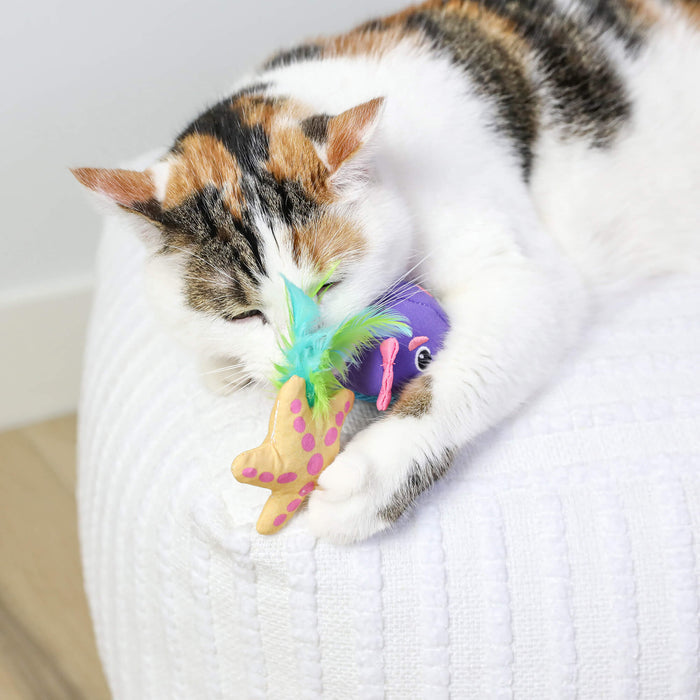 A calico cat lounges on a white cushion, playfully hugging and biting Quirky Kitty Pufferfish Pals Cat Toys 2-Pack—cooling catnip ocean toys by Quirky Kitty.