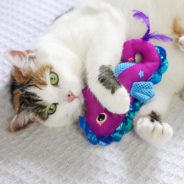 A white and brown cat with green eyes lies on a textured white surface, hugging the Quirky Kitty Sea Horse Kicker—a colorful ocean-themed catnip toy from Quirky Kitty—with its front paws.